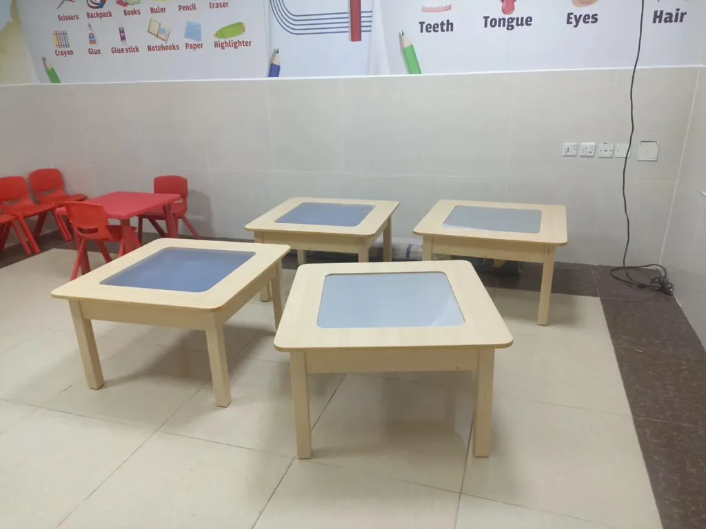 تجهيز الروضات:بطاولات تعليمية خشبية مربعة للأطفال في روضة مجهزة، بألوان فاتحة ومحيط تعليمي مزين برسومات أدوات الدراسة